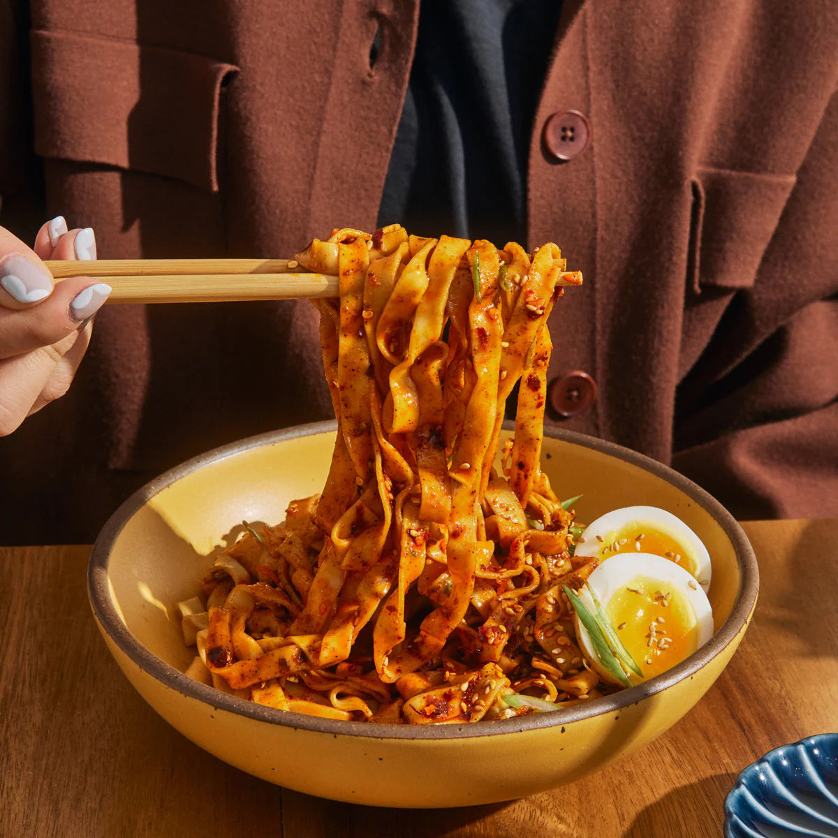 Chopsticks lifting chilli-coated noodles from a bowl with soft-boiled egg