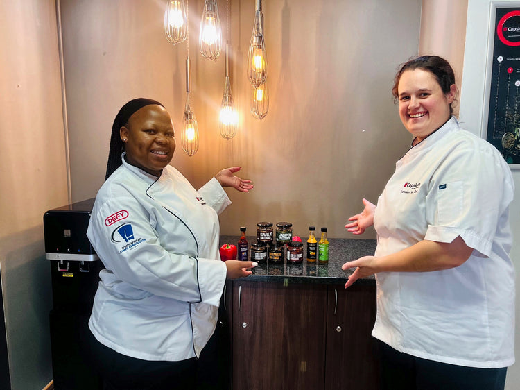 Two smiling chefs in whites presenting Kalahari Organics chilli crunch jars and hot sauces
